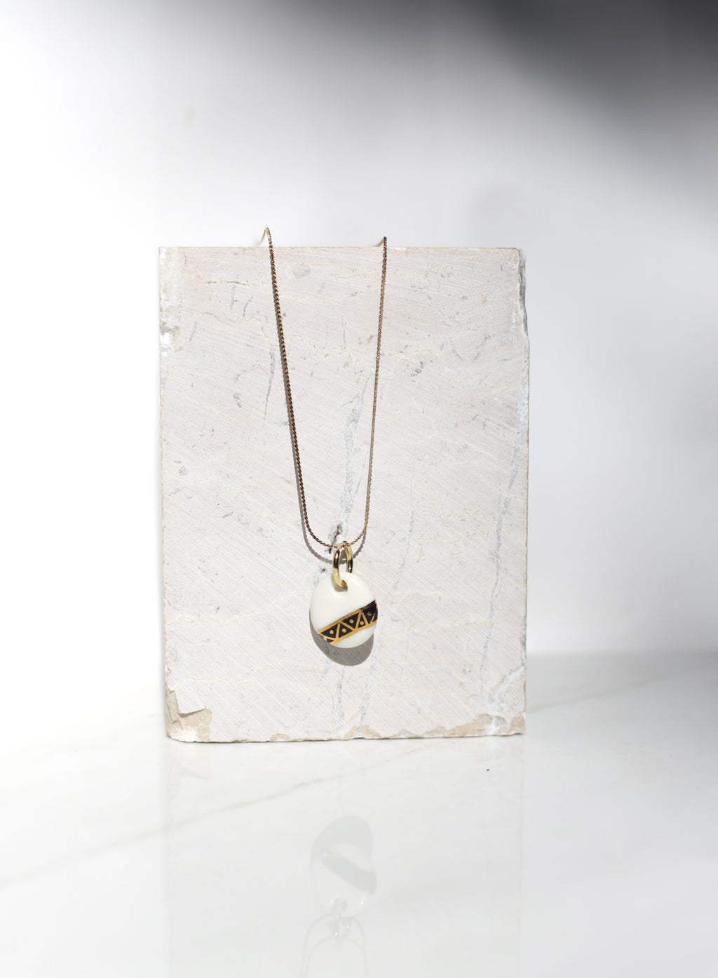 Collier composé d'une fine chaîne dorée et d'un pendentif circulaire en porcelaine blanche émaillée, orné d'un motif graphique transversale noire et or précieux aux motifs triangles géométriques de style Art Déco peint à la main 