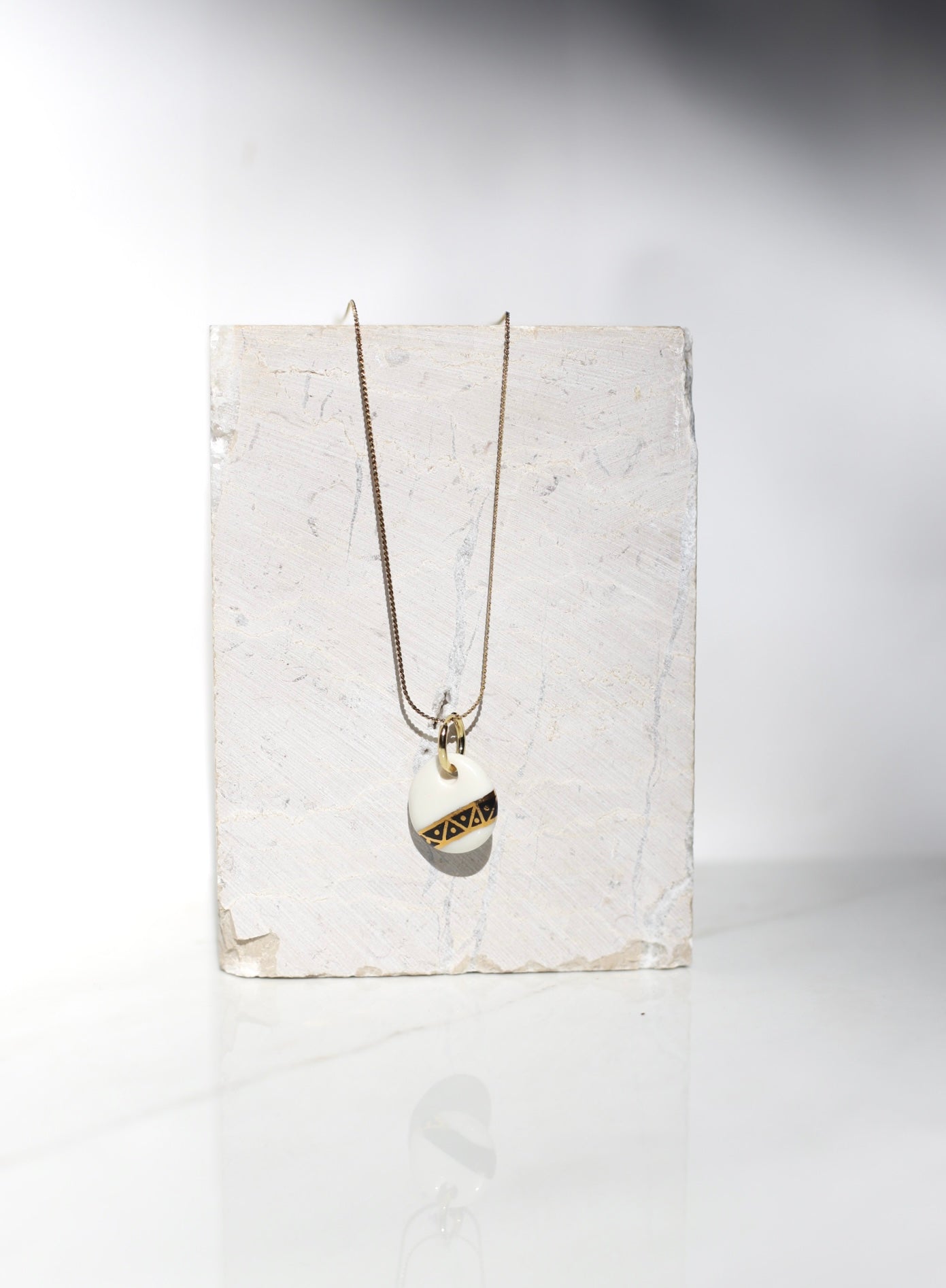 Collier composé d'une fine chaîne dorée et d'un pendentif circulaire en porcelaine blanche émaillée, orné d'un motif graphique transversale noire et or précieux aux motifs triangles géométriques de style Art Déco peint à la main 