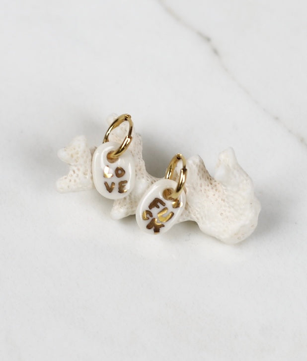 Paire de petites boucles d'oreilles pendantes asymétriques pour femme en porcelaine blanche, ornées des mots LOVE et FUCK écrits à l'or fin.