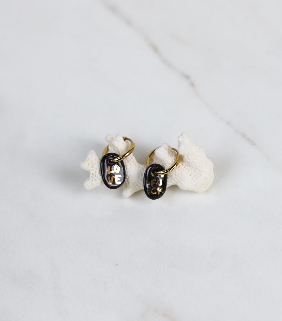 Paire de petites boucles d'oreilles pendantes asymétriques pour femme en porcelaine noire émaillée, ornées des mots LOVE et FUCK écrits en lettres dorées.