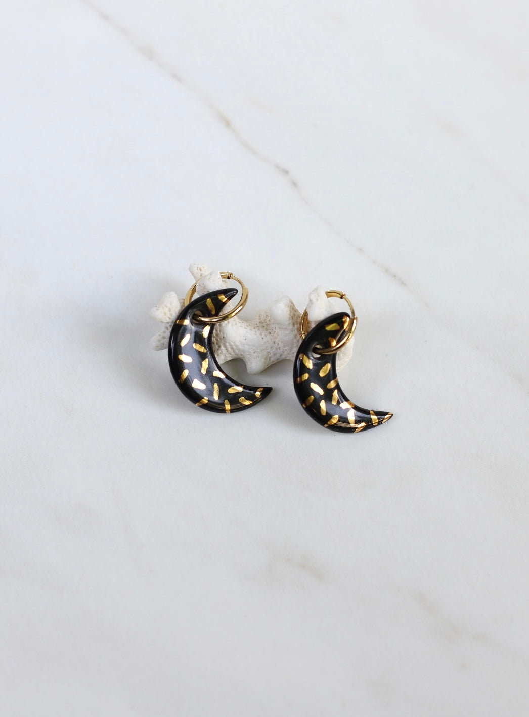 Une boucle d'oreille pendante dorée avec  en forme de croissant de lune noir brillant, décoré de petits traits dorés à l'or à la main. 