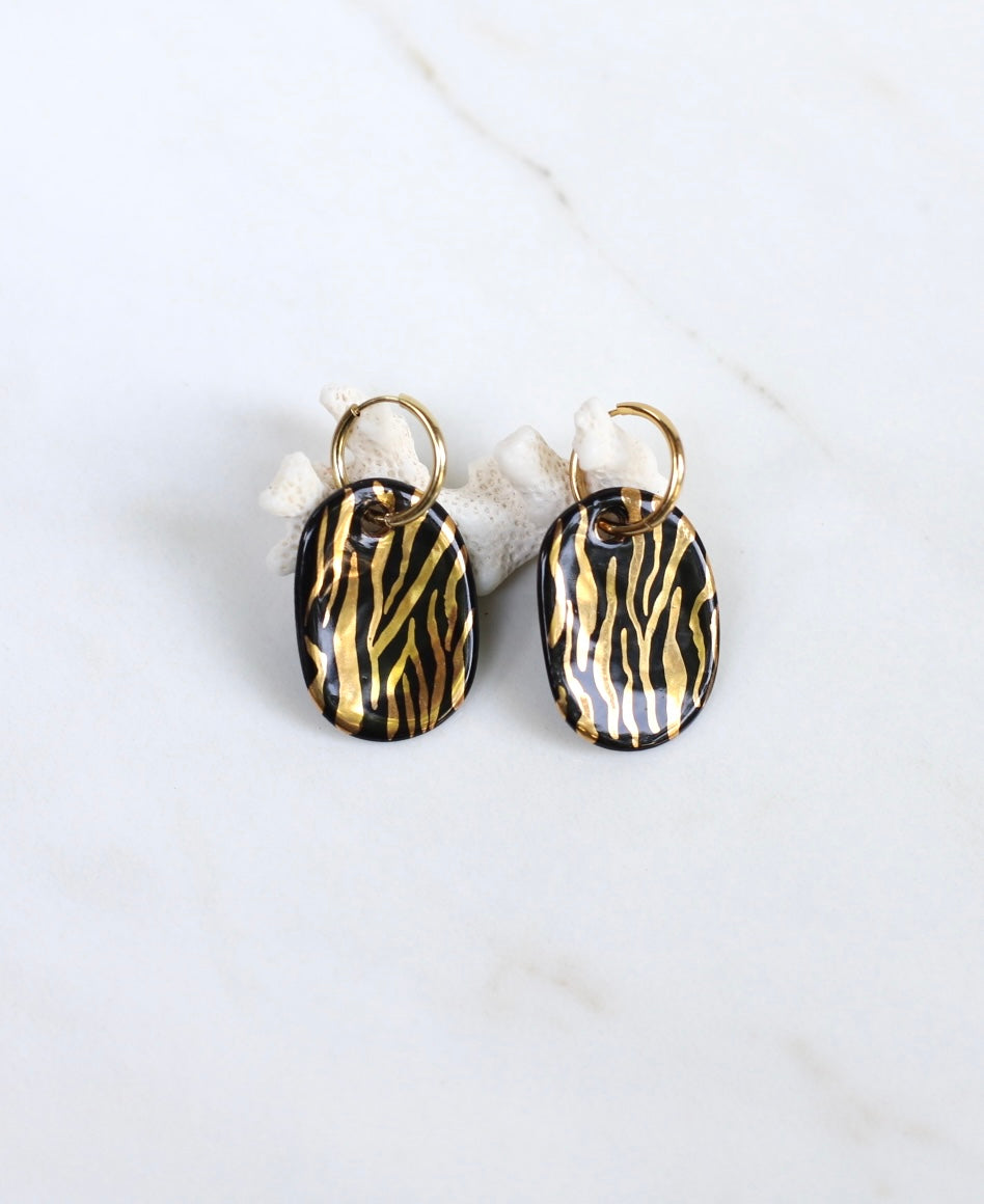 paire de boucles d'oreilles dorées avec des pendentifs de forme ovale en porcelaine noire ornés de rayures zébrées à l'or fin.