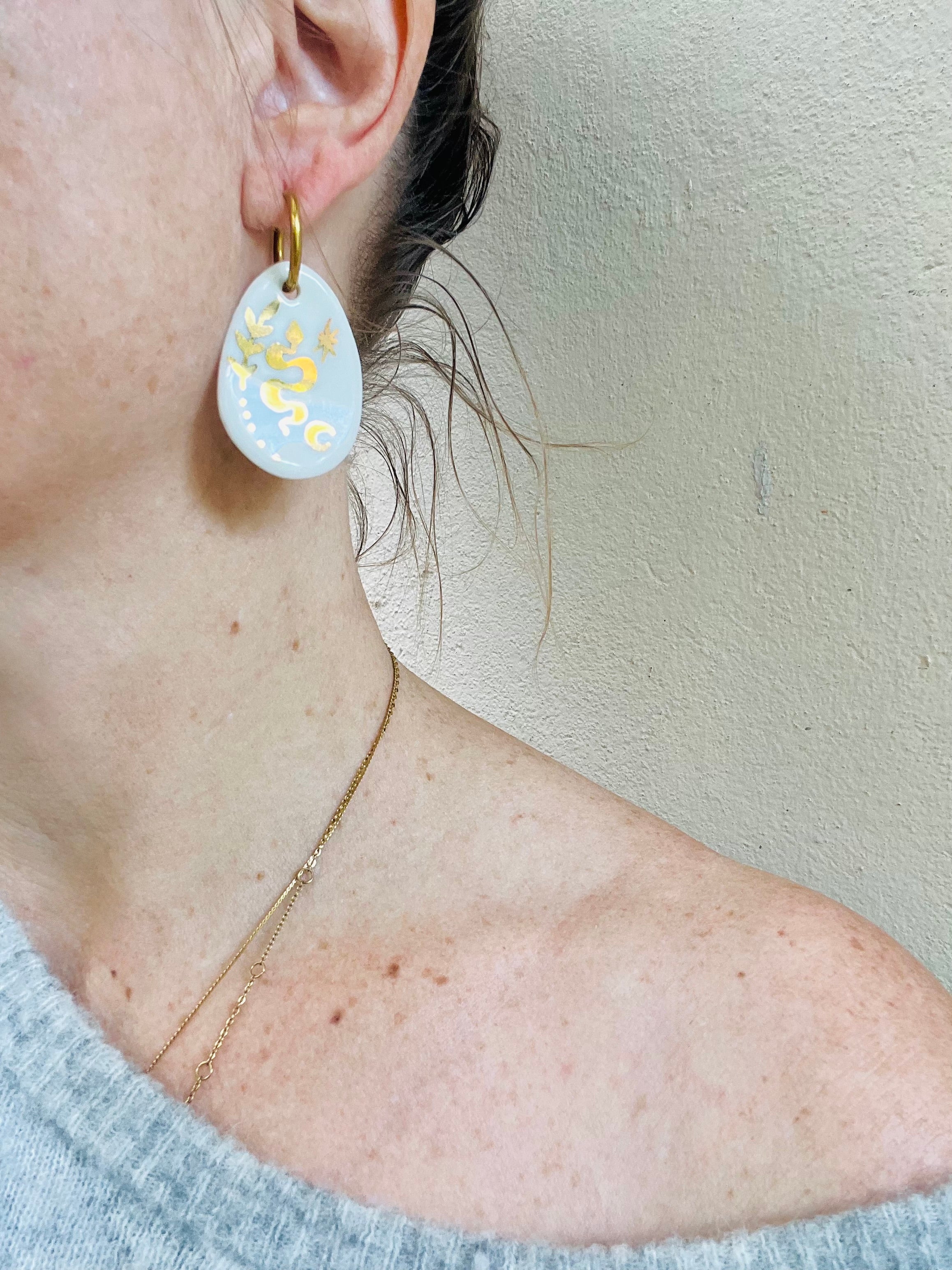 Boucle d'oreille mystique SNAKE portée par une femme. Boucles dorée avec motif en porcelaine blanche ornée d'un serpent et motifs célestes or peints à la main.