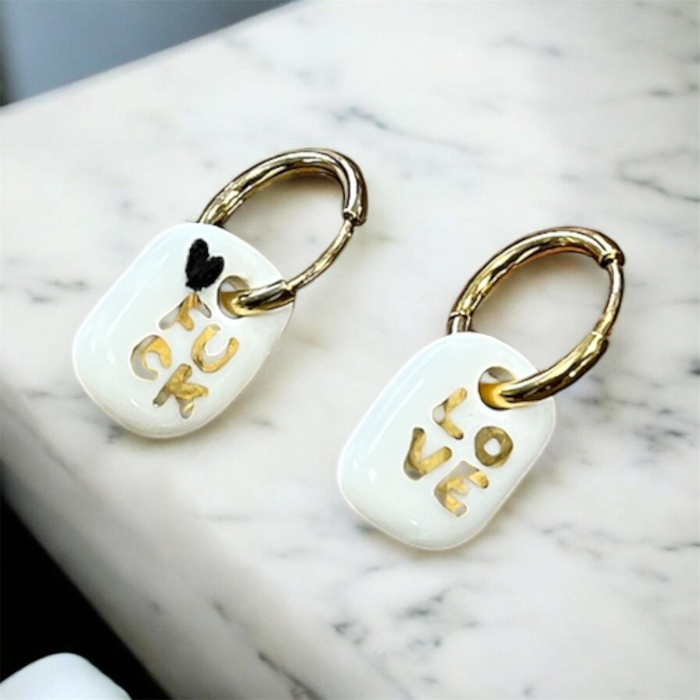 De boucles d'oreilles dépareillées en porcelaine blanche avec inscriptions dorées LOVE et FUCK et un petit détail de cœur noir