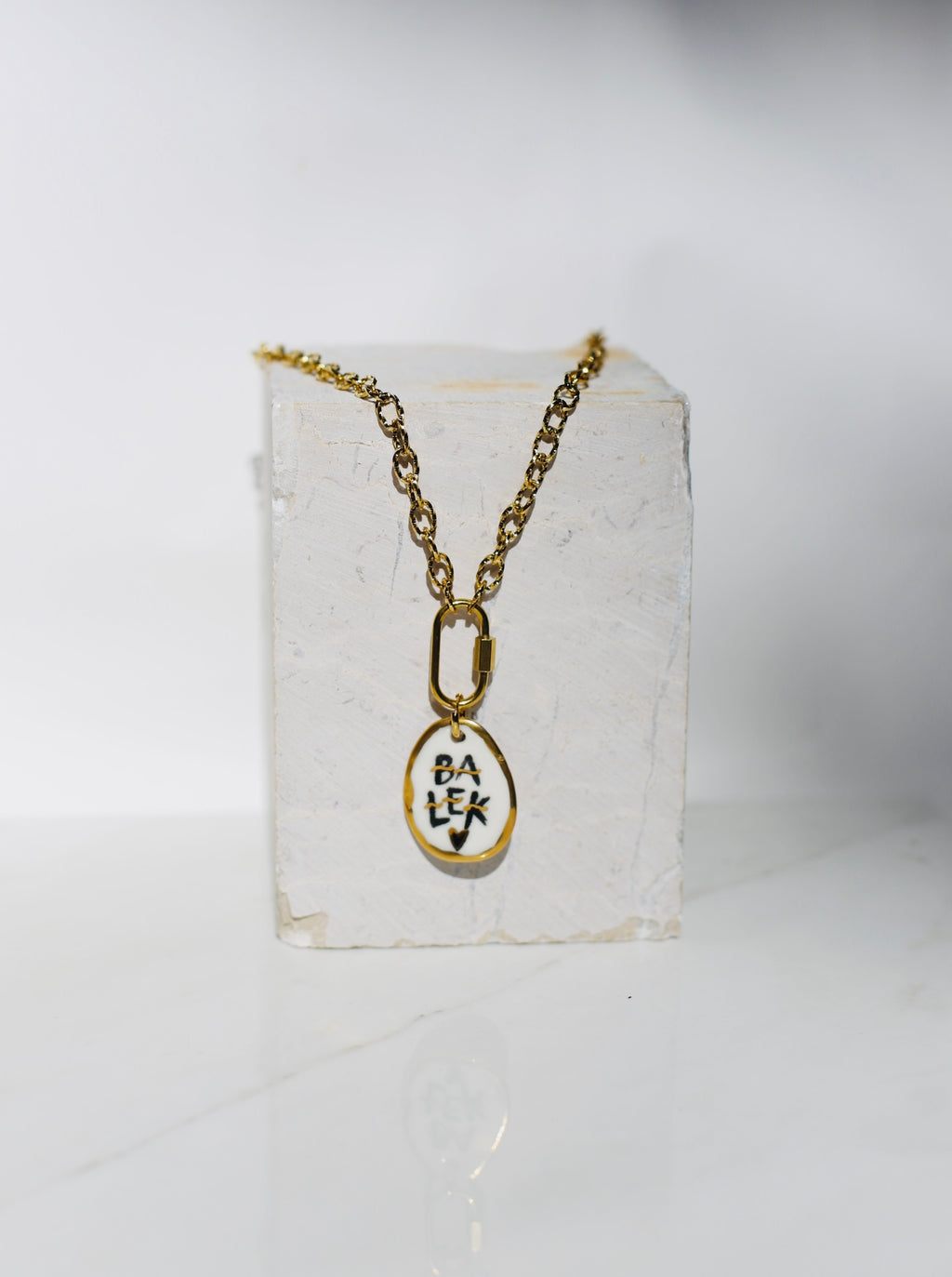 Chaine en acier inoxydable couleur or avec un médaillon en porcelaine montrant le mot BALEK écrit en noir et peint à la main avec des détails à l'or liquide 