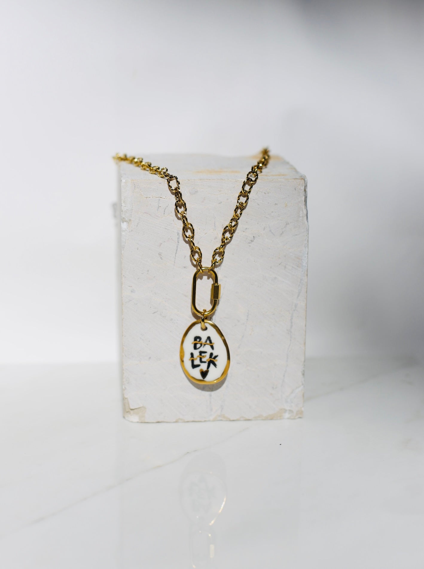 Chaine en acier inoxydable couleur or avec un médaillon en porcelaine montrant le mot BALEK écrit en noir et peint à la main avec des détails à l'or liquide 