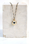 Collier romantique doté d'une chaîne doré perlée en porcelaine blanche émaillée, avec un médaillon où un  cœur noir est peint à la main avec des détails à l'or liquide 
