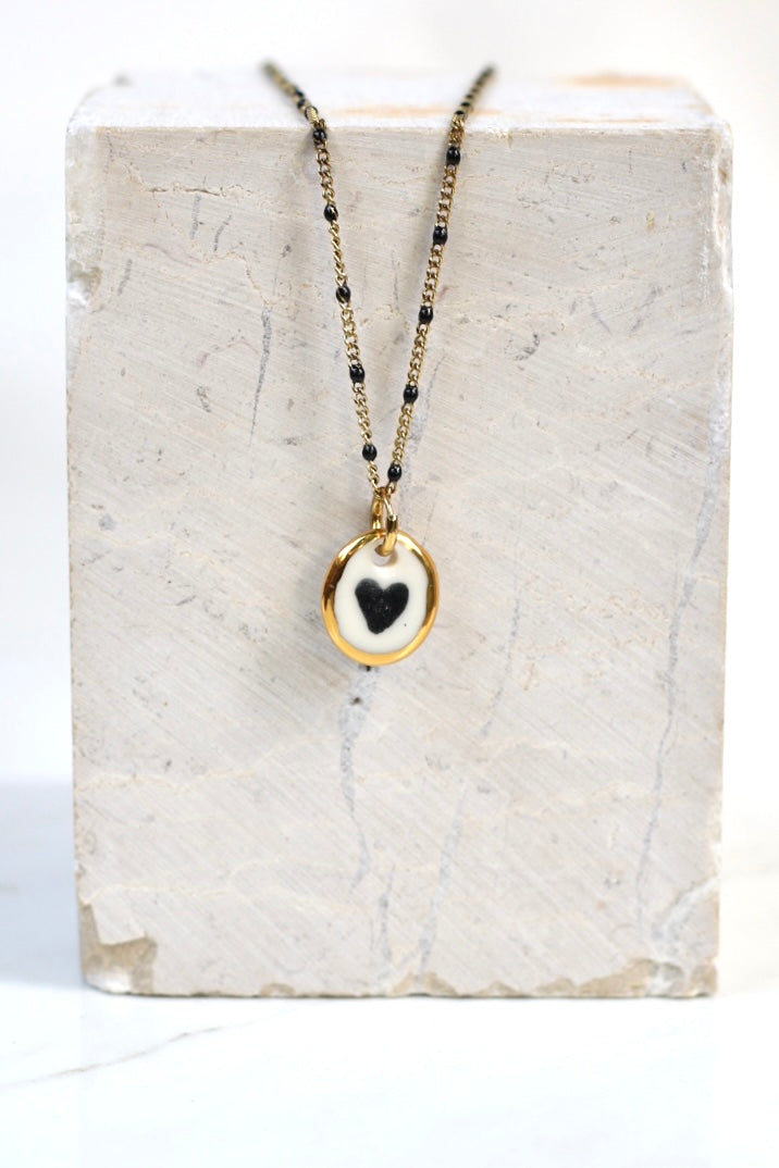 Collier romantique doté d'une chaîne doré perlée en porcelaine blanche émaillée, avec un médaillon où un  cœur noir est peint à la main avec des détails à l'or liquide 