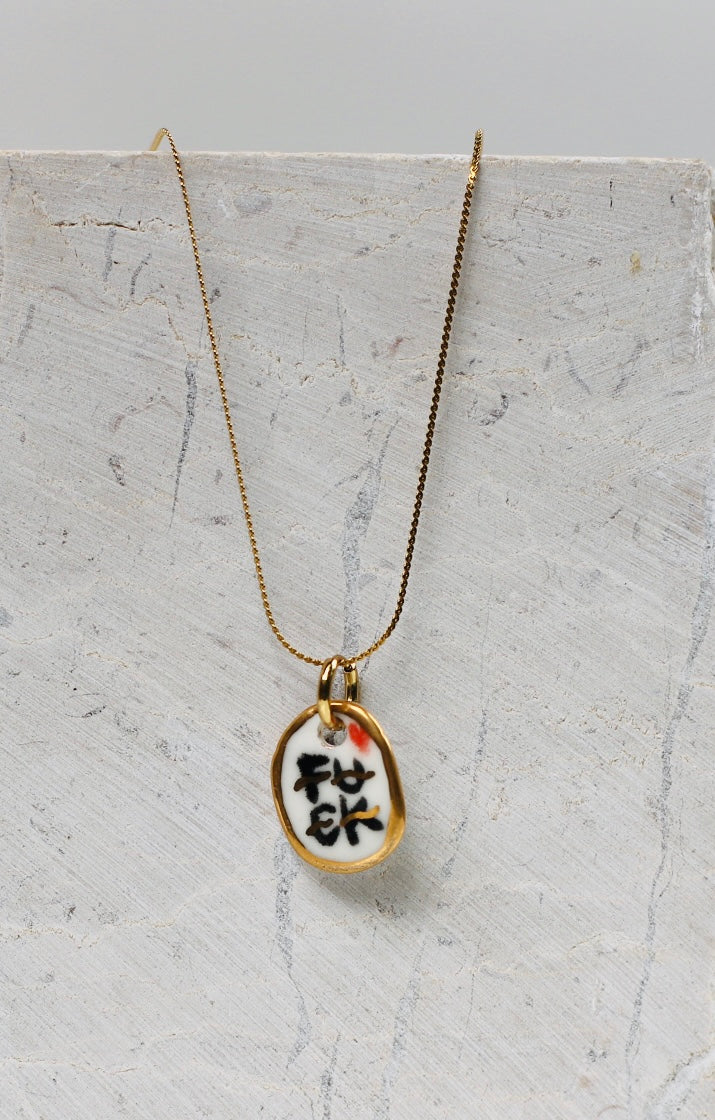 Collier pour femme avec un pendentif  ovale en porcelaine blanche. L'inscription FUCK est écrite en noir et soulignée d'or avec une bordure entièrement dorée