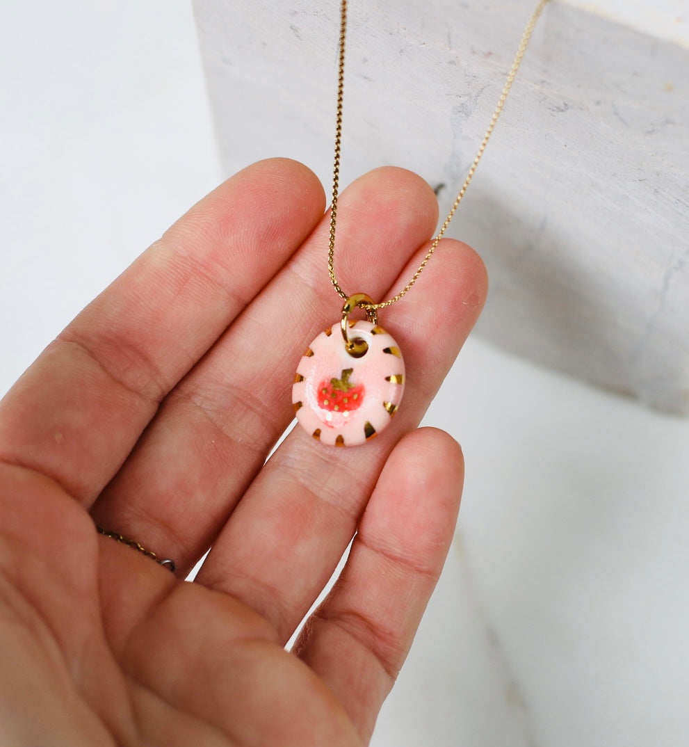 collier avec un pendentif rond en porcelaine émaillée rose représentant une petite fraise rouge aux pépins d'or et ses finitions artisanales.