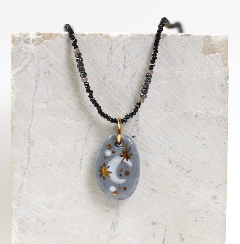 Collier pour femme avec un pendentif galet en porcelaine gris bleuté émaillée, orné d'un croissant de lune blanc et d'étoiles dorées à l'or fin, monté sur une chaîne de perles noires facettées.