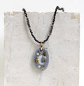 Collier pour femme avec un pendentif galet en porcelaine gris bleuté émaillée, orné d'un croissant de lune blanc et d'étoiles dorées à l'or fin, monté sur une chaîne de perles noires facettées.