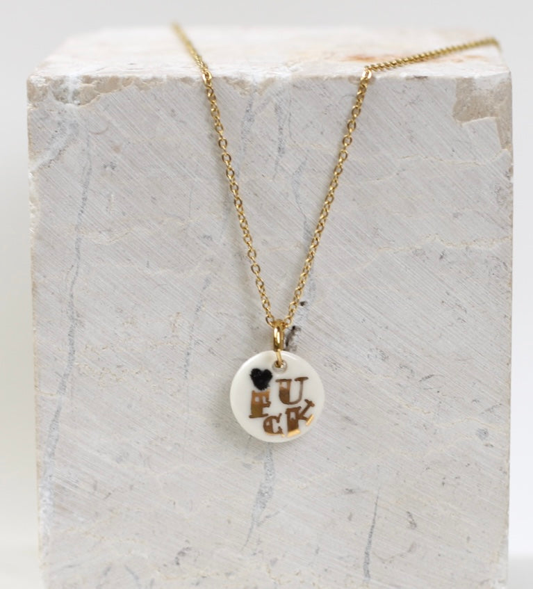 collier délicat composé d'une petite chaîne dorée et d'une médaille ronde en porcelaine blanche avec le mot FUCK écrit à l'or fin et un mini cœur noir.