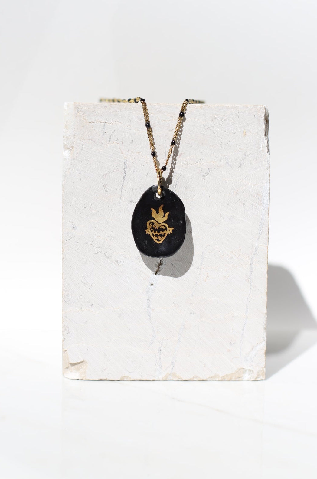 Collier en porcelaine noire et or fin au style rock chic, mettant en valeur la forme irrégulière du galet et la brillance du sacré-cœur ex-voto.