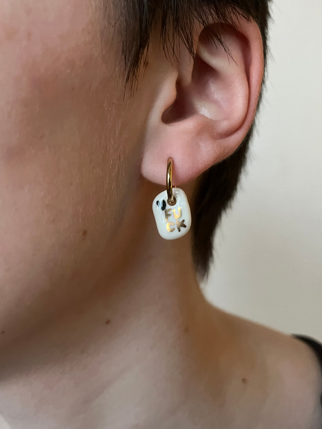 Boucle d'oreille rock en porcelaine blanche et or portée à l'oreille, affichant le message FUCK peint à la main à l'or liquide avec un mini cœur noir