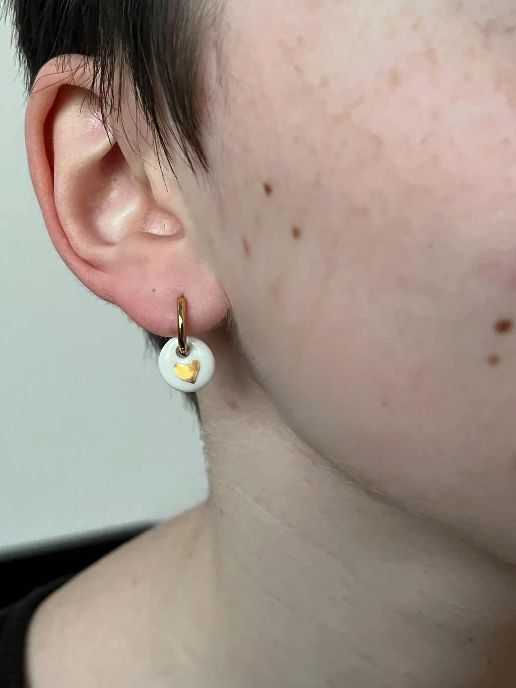 Petite boucle d'oreille pendante minimaliste en porcelaine ornée d'un cœur doré, portée à l'oreille pour un style discret et élégant au quotidien.
