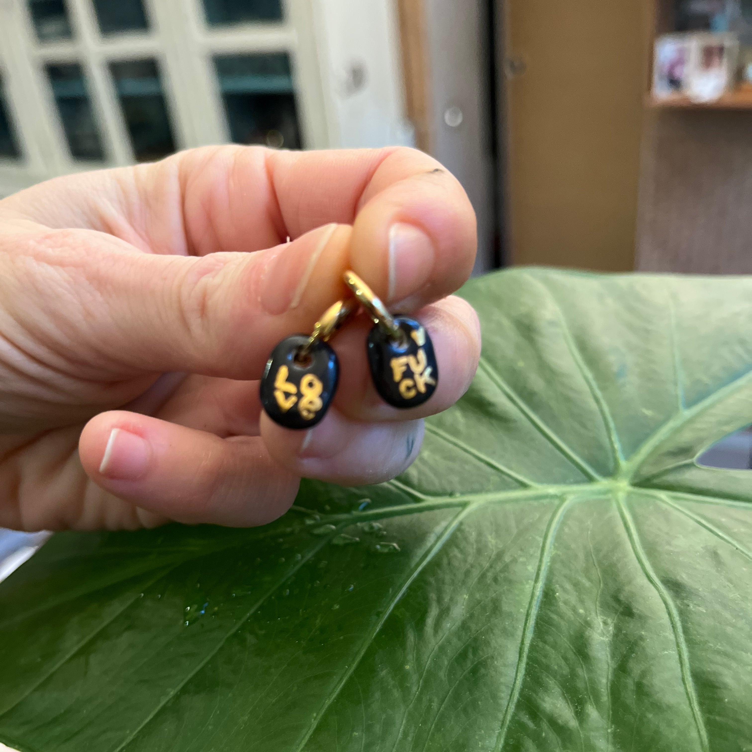 Gros plan de deux boucles d'oreilles à message en porcelaine noire et lettres dorées, tenues entre les doigts pour montrer les inscriptions LOVE et FUCK.