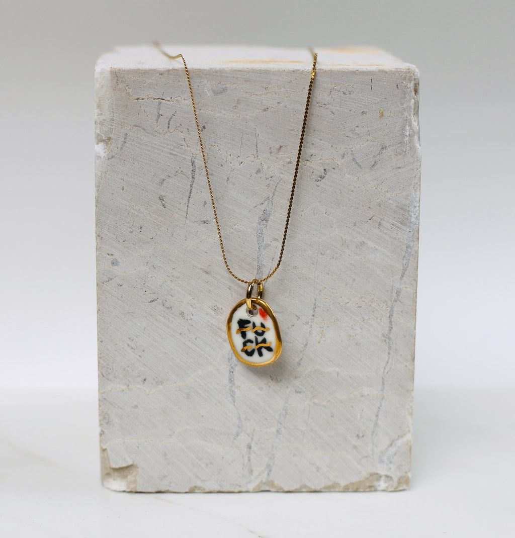 collier avec pendentif ovale irrégulier en porcelaine émaillée blanche, affichant le mot FUCK écrit dans un style calligraphique noir et doré à l'or liquide.