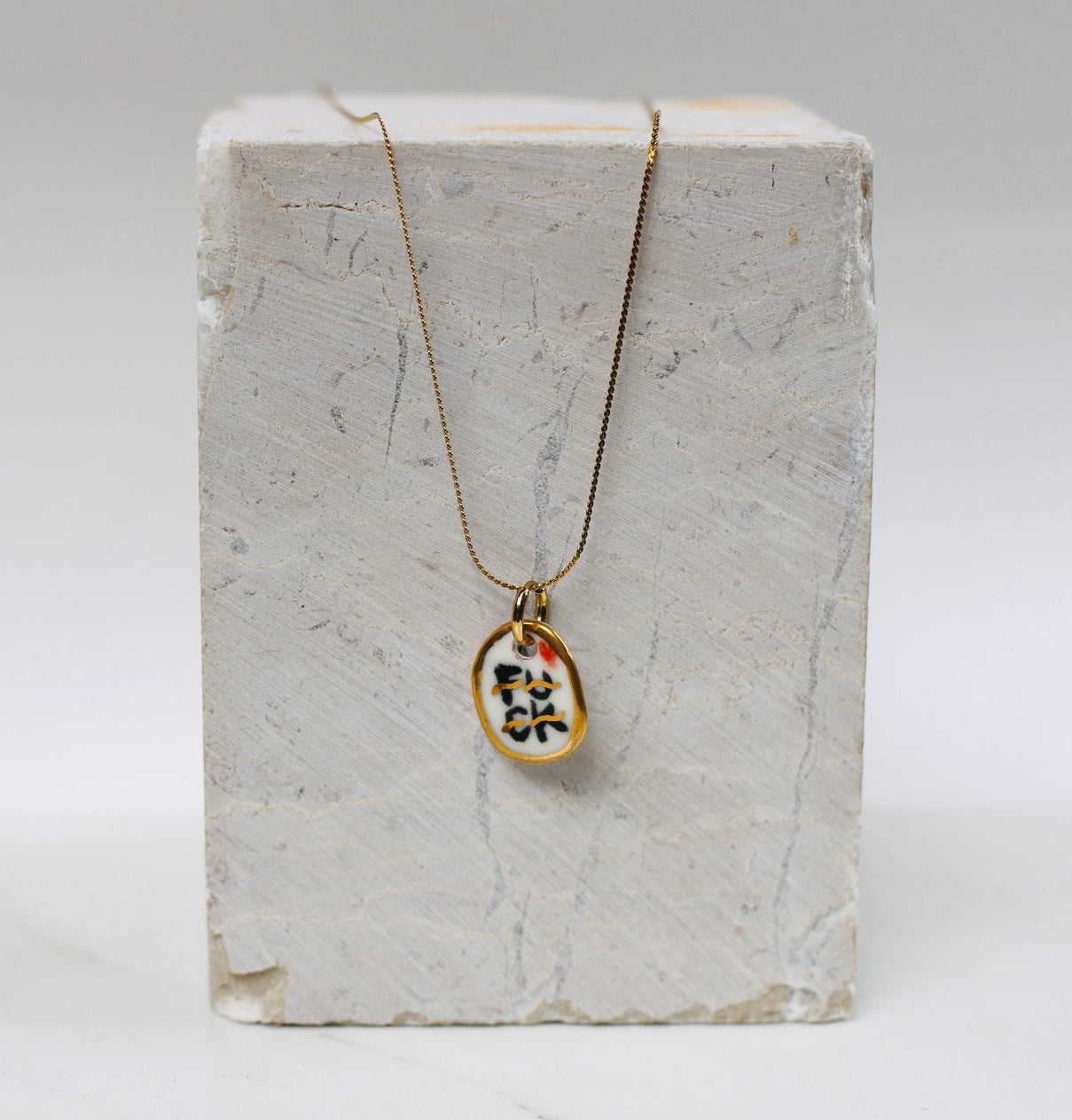 collier avec pendentif ovale irrégulier en porcelaine émaillée blanche, affichant le mot FUCK écrit dans un style calligraphique noir et doré à l'or liquide.