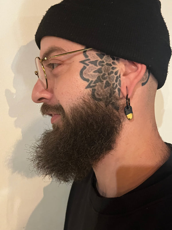 Boucle d'oreille noir avec un petit pendentif en porcelaine noire et pointe d'or, portée de profil par un homme barbu arborant un tatouage sur la tempe et un bonnet noir.