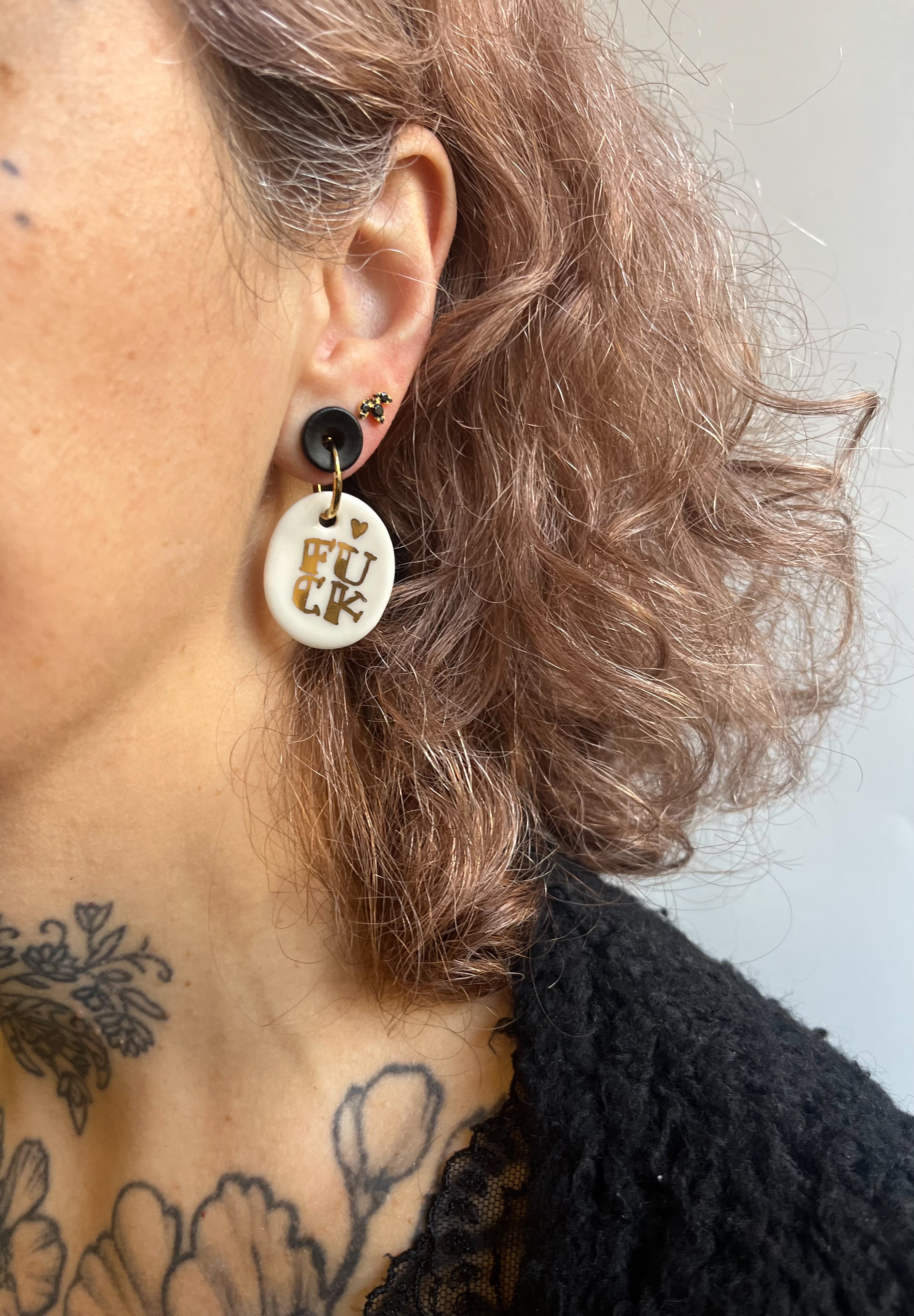 boucle d'oreille dorée avec son pendentif en porcelaine blanche, affichant le mot FUCK écrit en lettres dorées à l'or fin.
