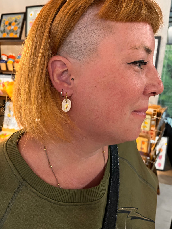 Boucle d'oreille dorée Matilou avec un petit pendentif en porcelaine blanche, délicatement orné d'un motif de feuille dorée à l'or. bijou porté en gros plan de profil par une cliente au style rock affirmé, avec des cheveux orange vif rasés sur le côté et un piercing au nez.