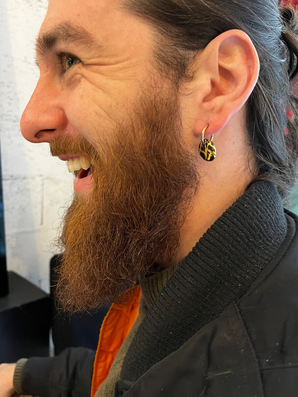 Petite boucle d'oreille anneau doré avec pendentif en porcelaine noire et motifs en or, portée de profil par un homme barbu au style rock