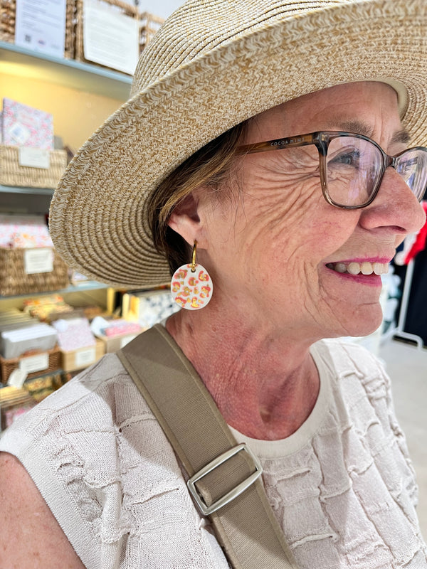 Boucle d'oreille dorée avec un grand pendentif rond en porcelaine blanche orné d'un motif léopard rose et d'éclats d'or pur, portée de profil par une cliente souriante avec un chapeau de paille et des lunettes.