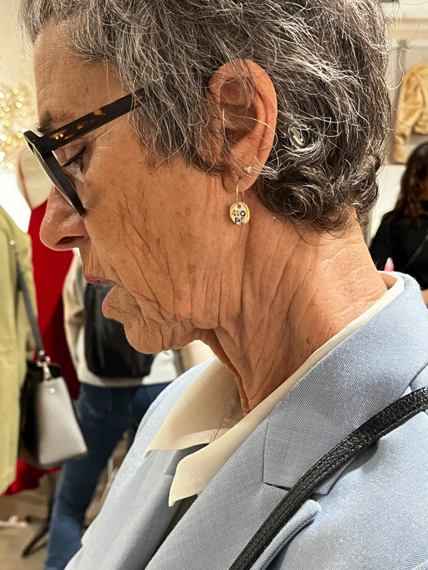 Petite boucle d'oreille ronde en porcelaine blanche Matilou avec lettres de style tatouage "HOPE" et petite étoile en or, portée par une cliente élégante aux cheveux poivre et sel et veste bleue.