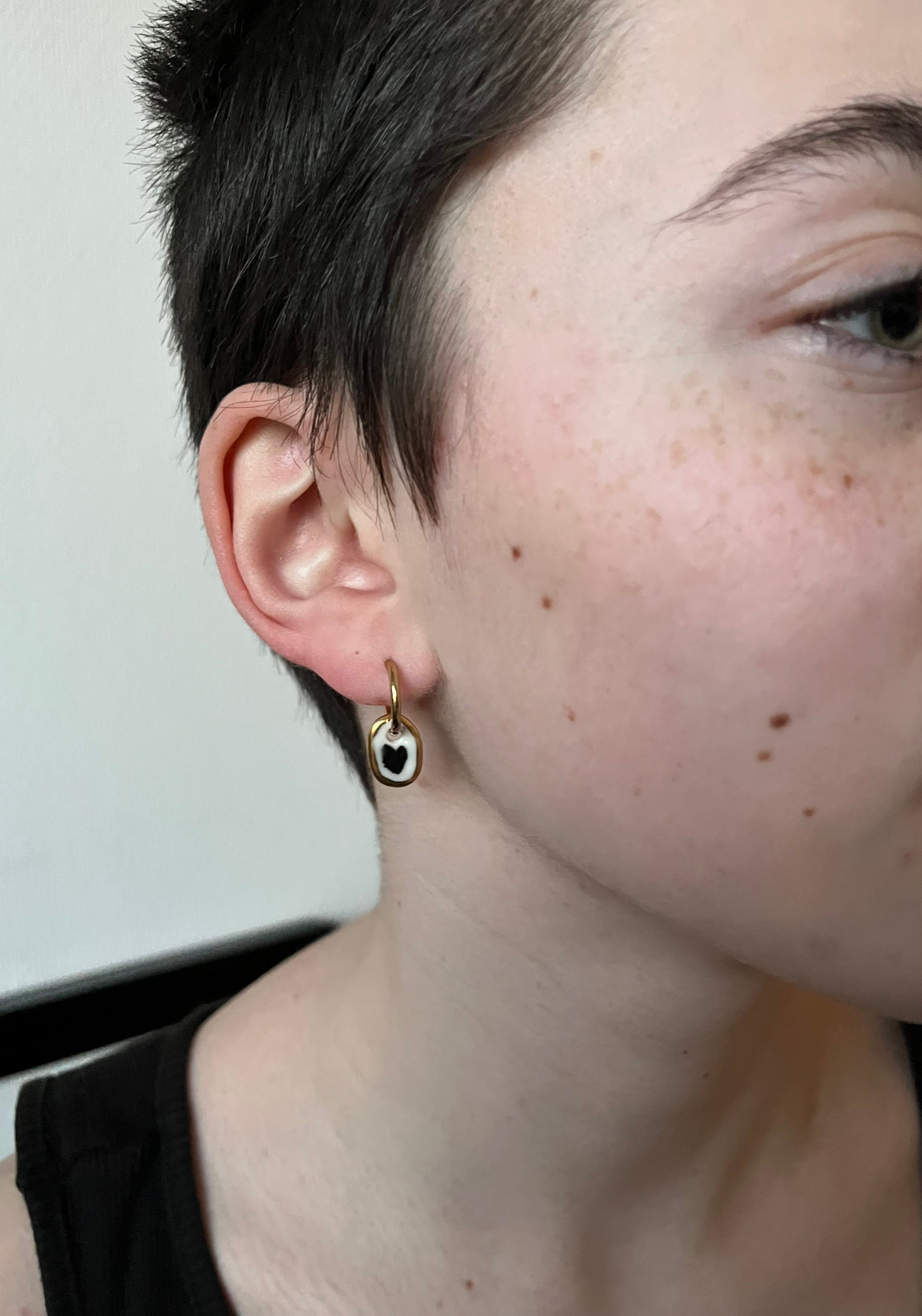 boucle d'oreille pendante avec un anneau doré en porcelaine blanc orné d'un cœur noir peint à la main et d'un contour doré.