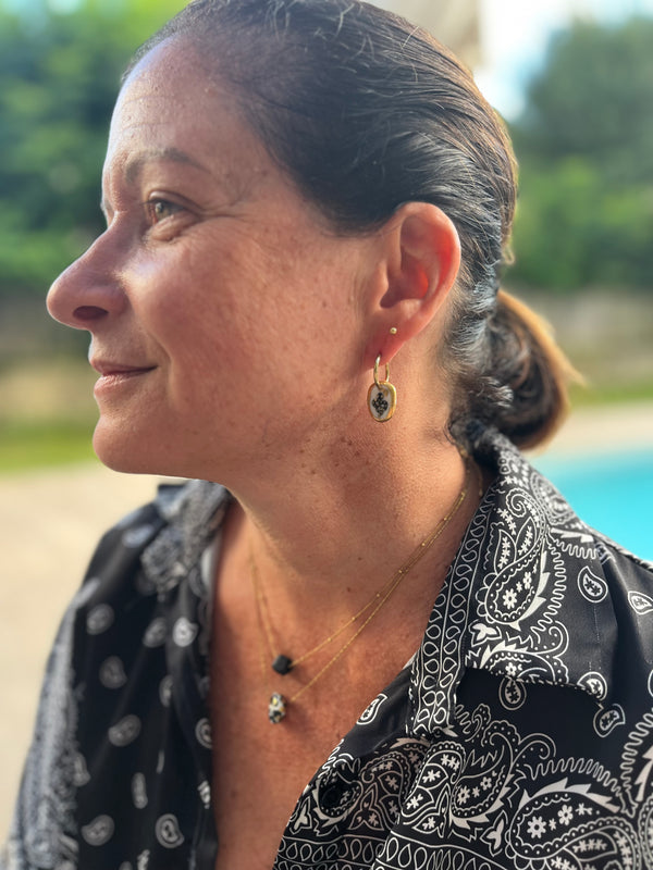Boucle d'oreille dorée avec petit pendentif ovale en porcelaine blanche, illustration noire et bordure en or, portée de profil avec une accumulation de colliers fins Matilou et une chemise bandana.