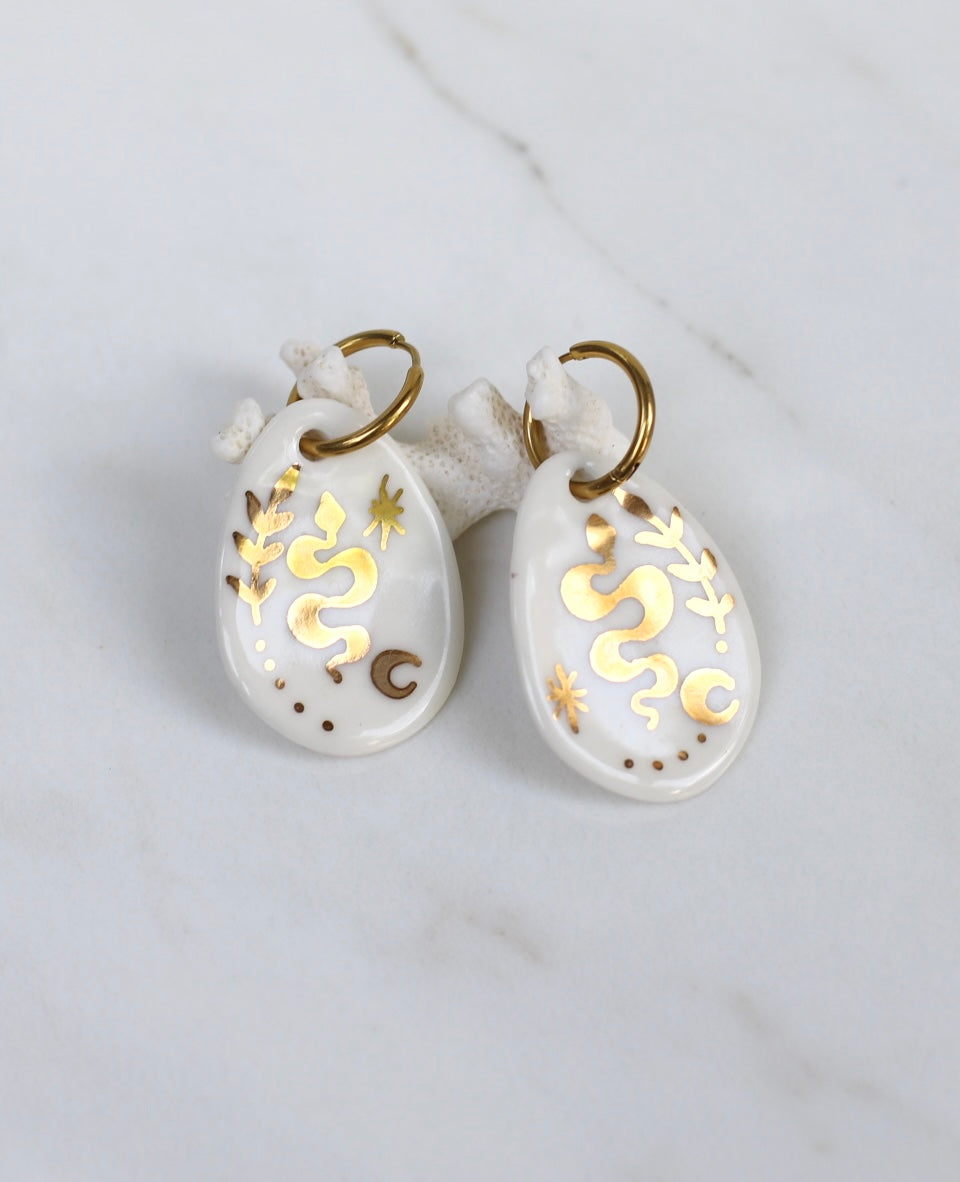 Paire de boucles d'oreilles pendantes symboliques en porcelaine blanche et or fin. Motif serpent sinueux, étoile et lune peints à la main 