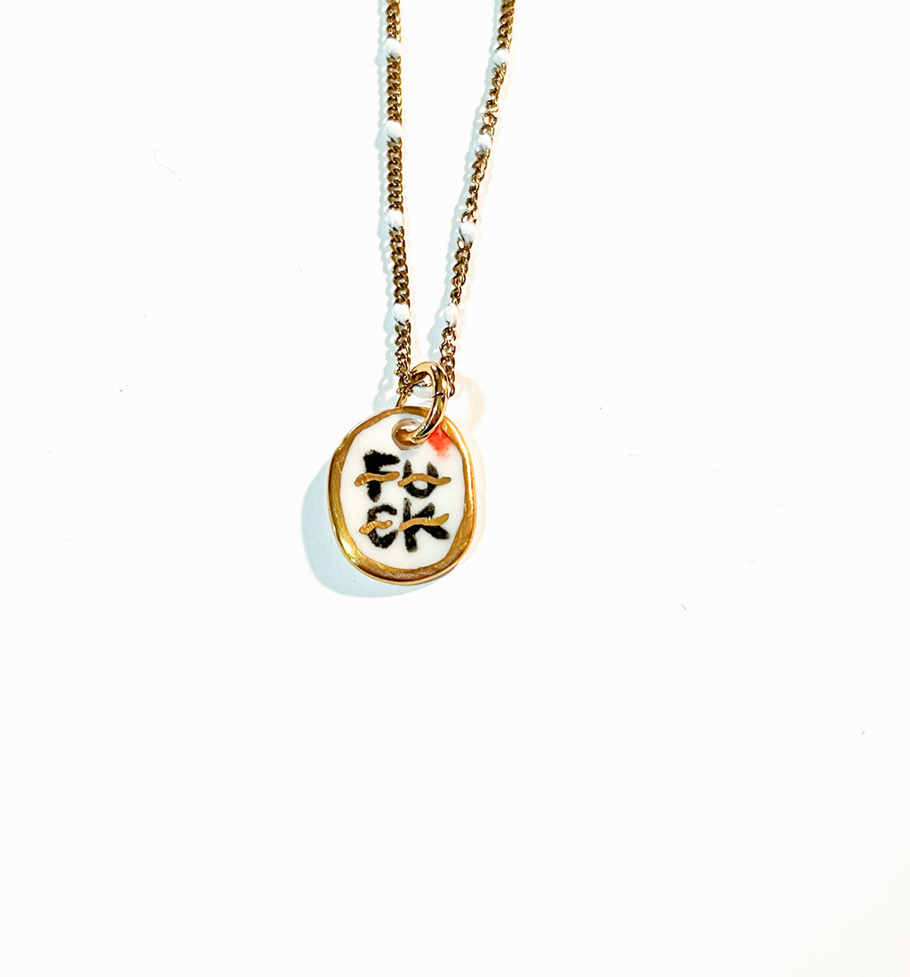 collier pendentif en porcelaine blanche avec le message rock FUCK écrit en noir et or, monté sur une fine chaîne dorée.