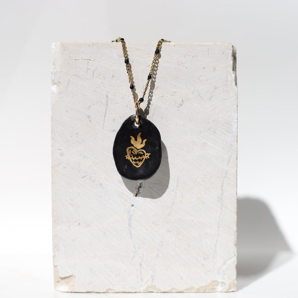 Collier avec un pendentif galet en porcelaine noire émaillée orné d'un dessin de sacré-cœur ex-voto enflammé à l'or fin