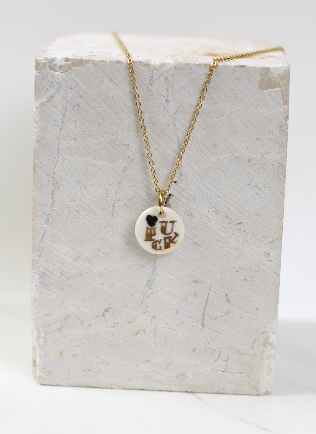 Pendentif artisanal en porcelaine émaillée blanche portant l'inscription FUCK dorée à l'or liquide, monté sur une chaîne dorée scintillante fais main