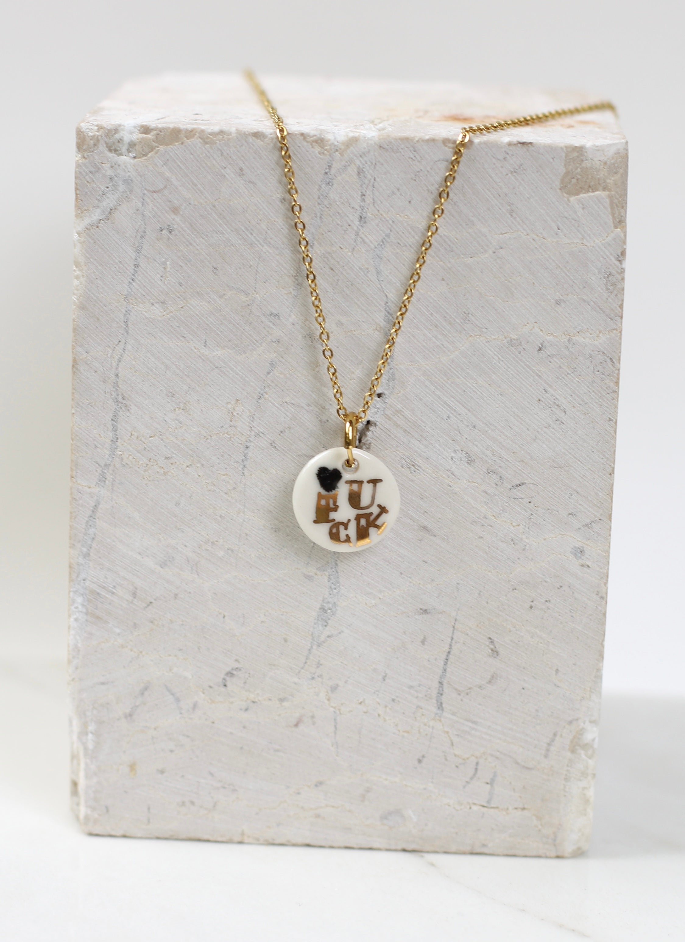 Pendentif artisanal en porcelaine émaillée blanche portant l'inscription FUCK dorée à l'or liquide, monté sur une chaîne dorée scintillante fais main