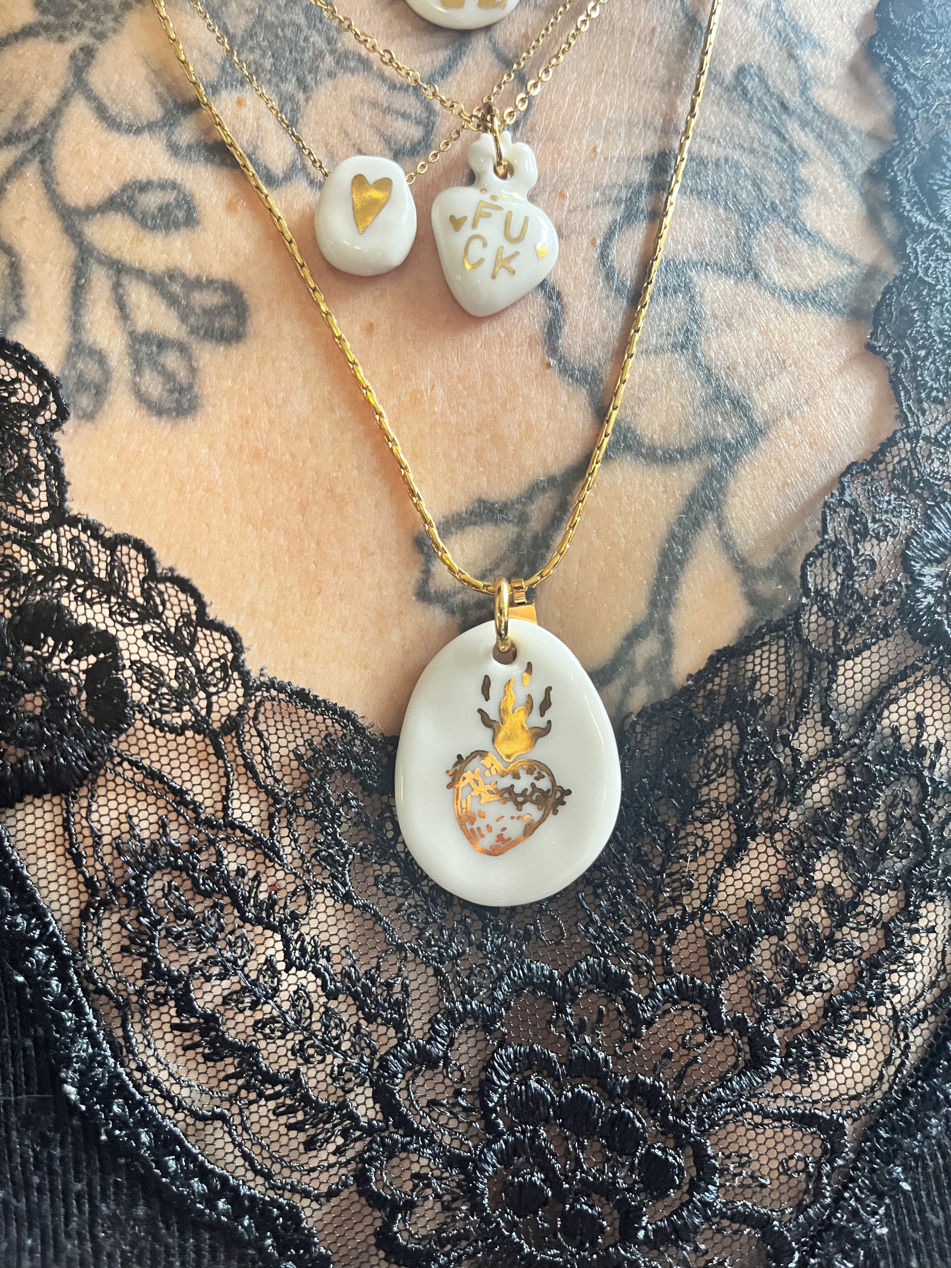 Gros plan d'un collier en porcelaine blanche et or mettant en valeur le grand pendentif galet et son motif cœur ex-voto doré.