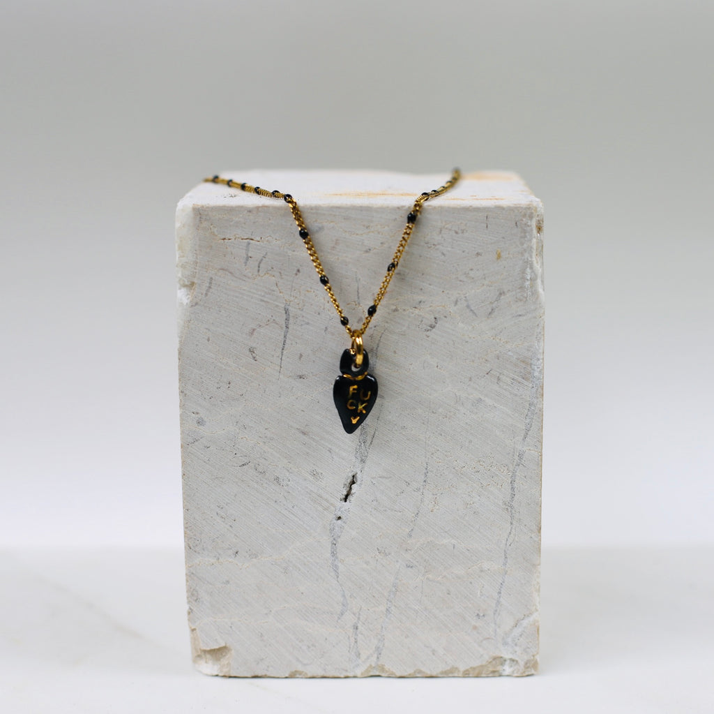 Collier pour femme avec une fine chaîne dorée à perles noires et un petit pendentif en porcelaine noire émaillée en forme de cœur ex-voto avec le mot FUCK écrit à l'or fin