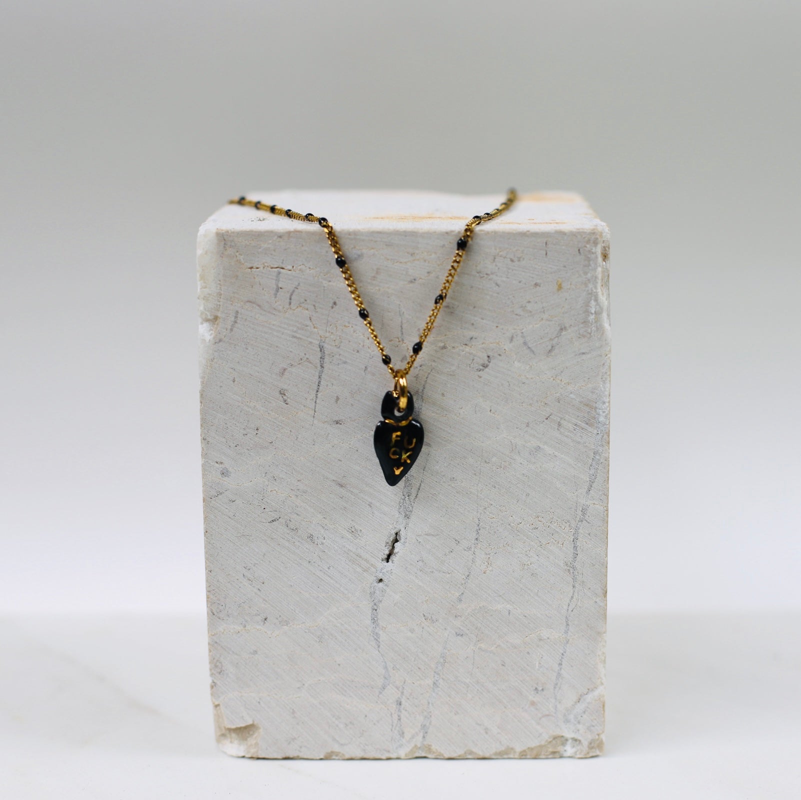 Collier pour femme avec une fine chaîne dorée à perles noires et un petit pendentif en porcelaine noire émaillée en forme de cœur ex-voto avec le mot FUCK écrit à l'or fin