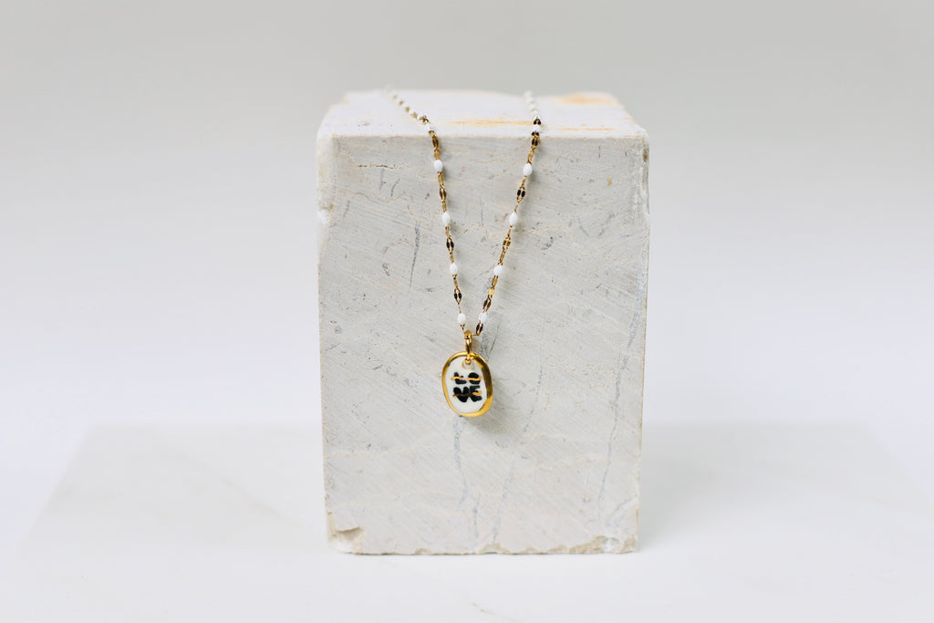 Collier pour femme avec un pendentif galet ovale en porcelaine blanche. Le mot LOVE est écrit en noir avec des traits d'or et une bordure dorée, monté sur une chaîne perlée blanche et dorée.