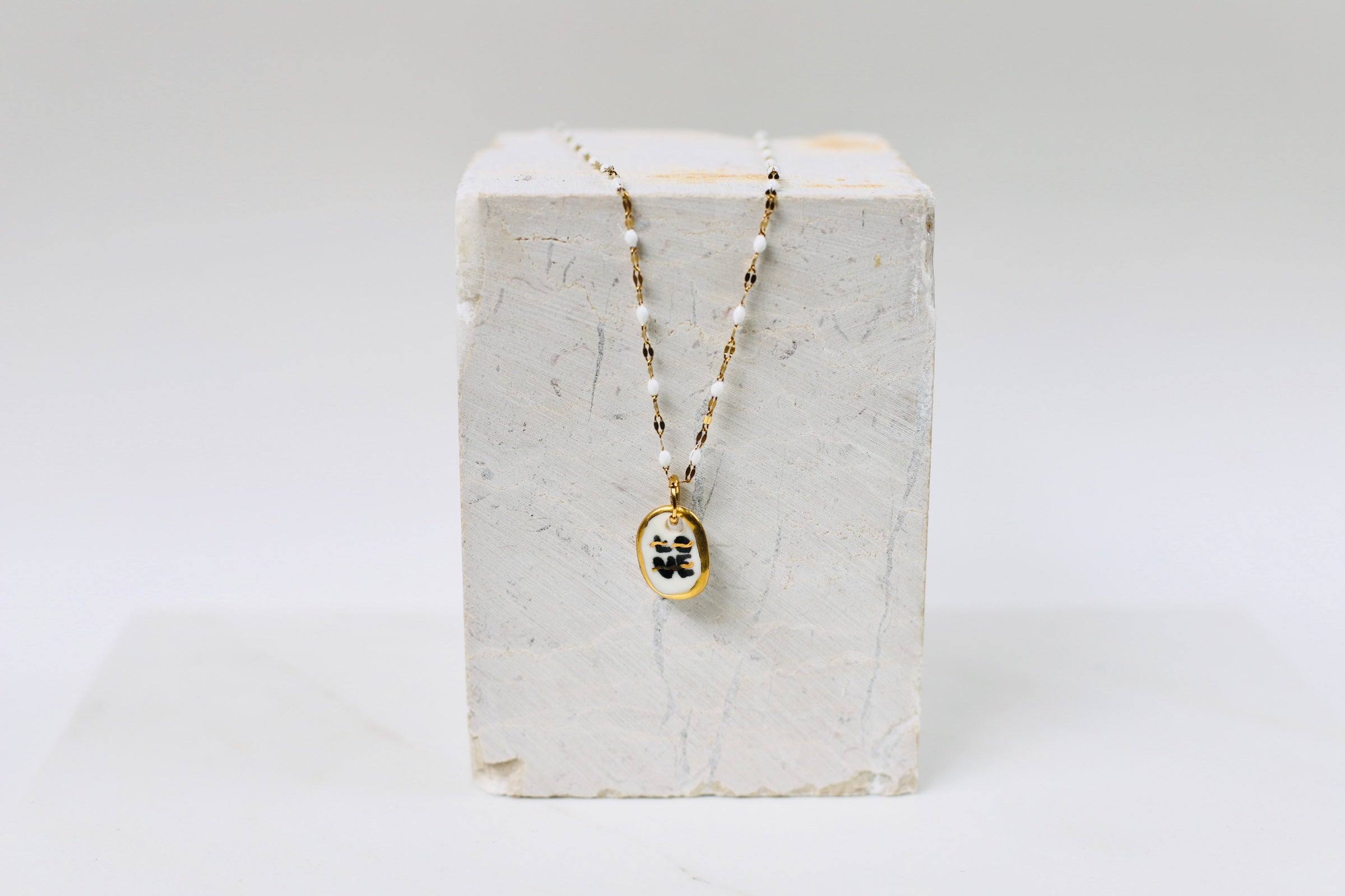 Collier pour femme avec un pendentif galet ovale en porcelaine blanche. Le mot LOVE est écrit en noir avec des traits d'or et une bordure dorée, monté sur une chaîne perlée blanche et dorée.