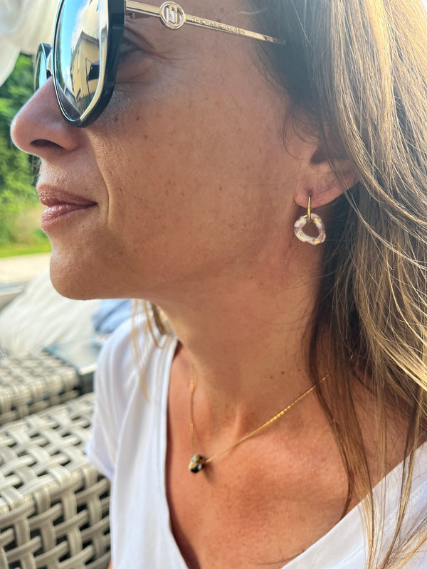 Boucle d'oreille créole dorée avec un pendentif anneau organique en porcelaine blanche et éclats d'or, portée en duo avec un collier fin Matilou par une cliente de profil.