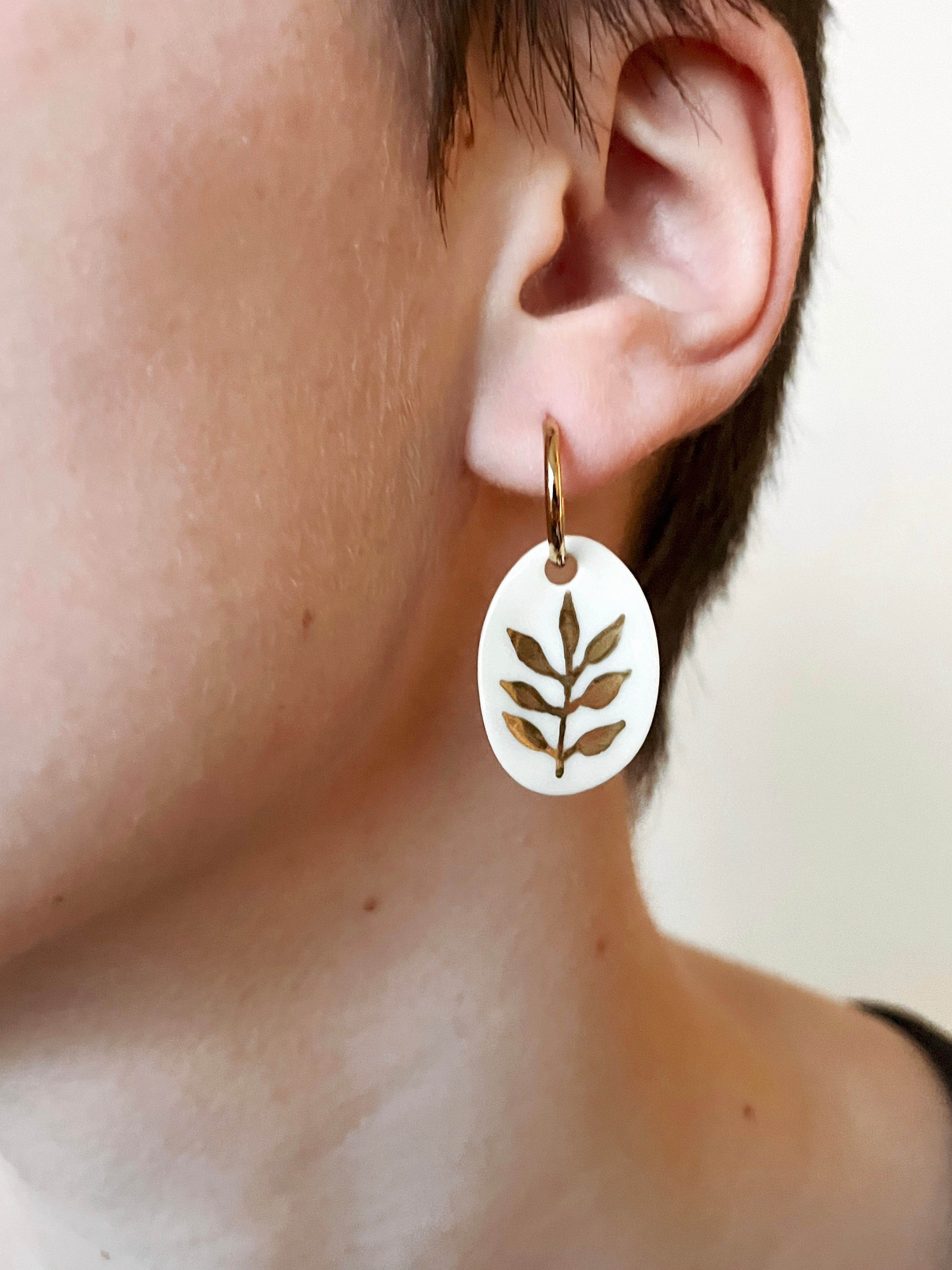 Boucle d'oreille  ovale en porcelaine et or portée par une femme. Bijou artisanal délicat avec motif floral doré, idéal pour un style élégant et poétique
