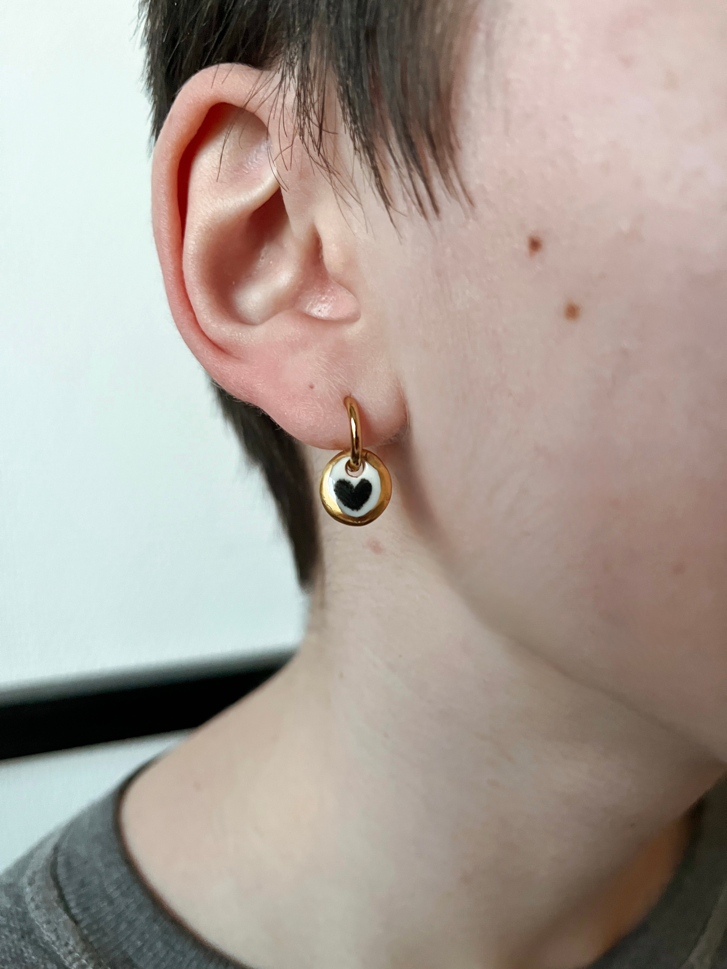 boucle d'oreille dorée avec petite médaille  en porcelaine blanche, avec un motif cœur noir entouré d'un liseré d'or fait à la main 