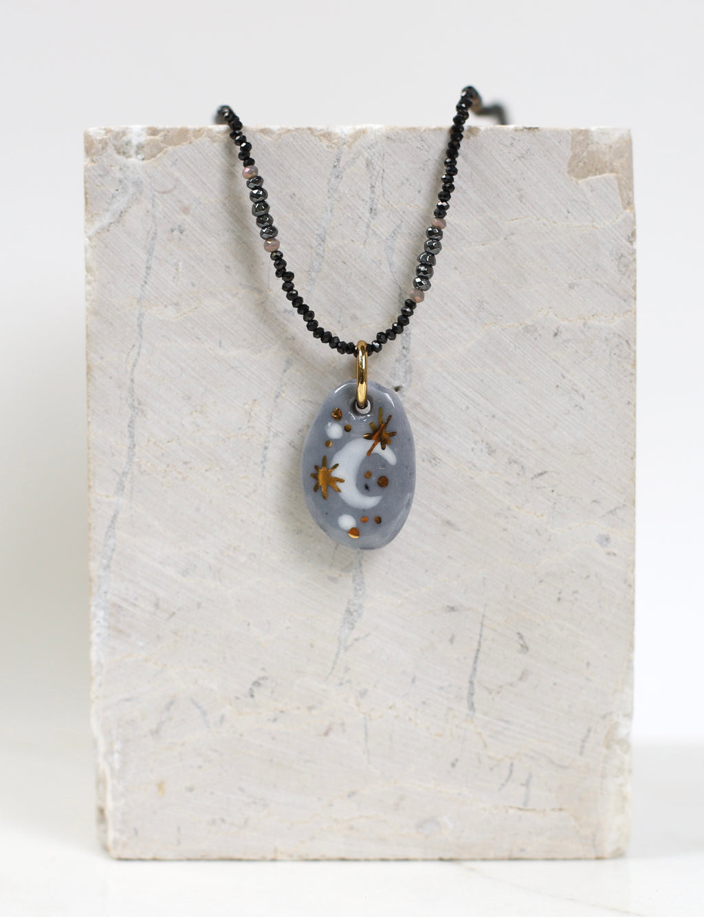 collier astral en porcelaine grise et or fin, mettant en valeur le motif lune peint à la main sur le médaillon et le détail de la fine chaîne perlée sombre.