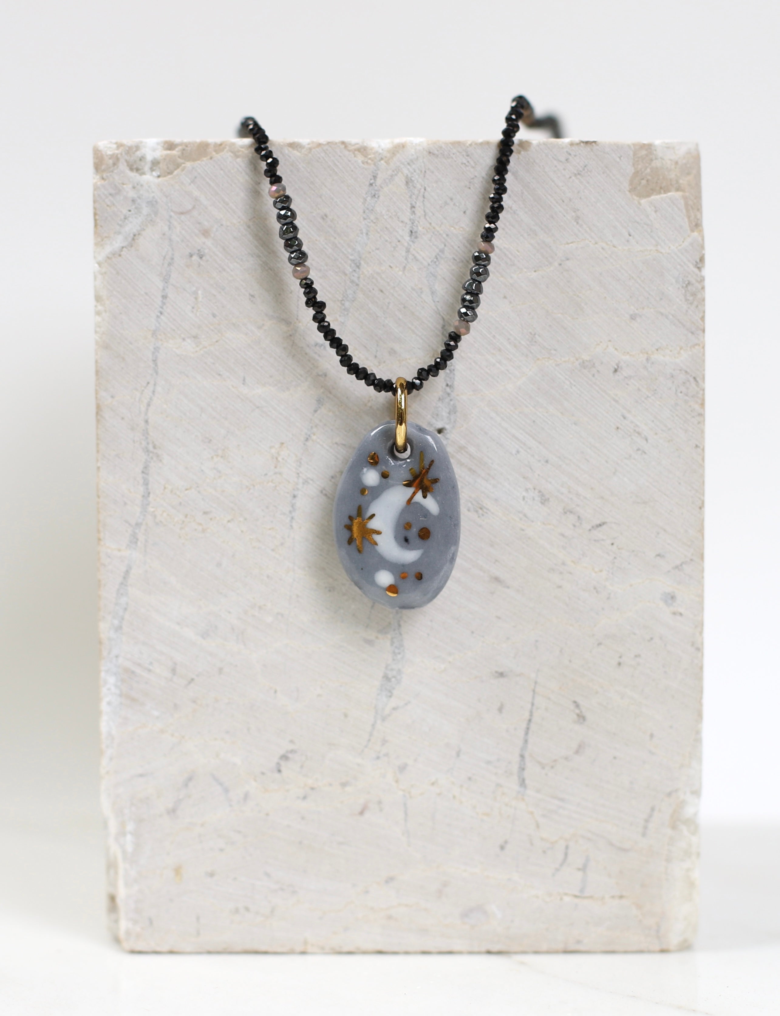collier astral en porcelaine grise et or fin, mettant en valeur le motif lune peint à la main sur le médaillon et le détail de la fine chaîne perlée sombre.