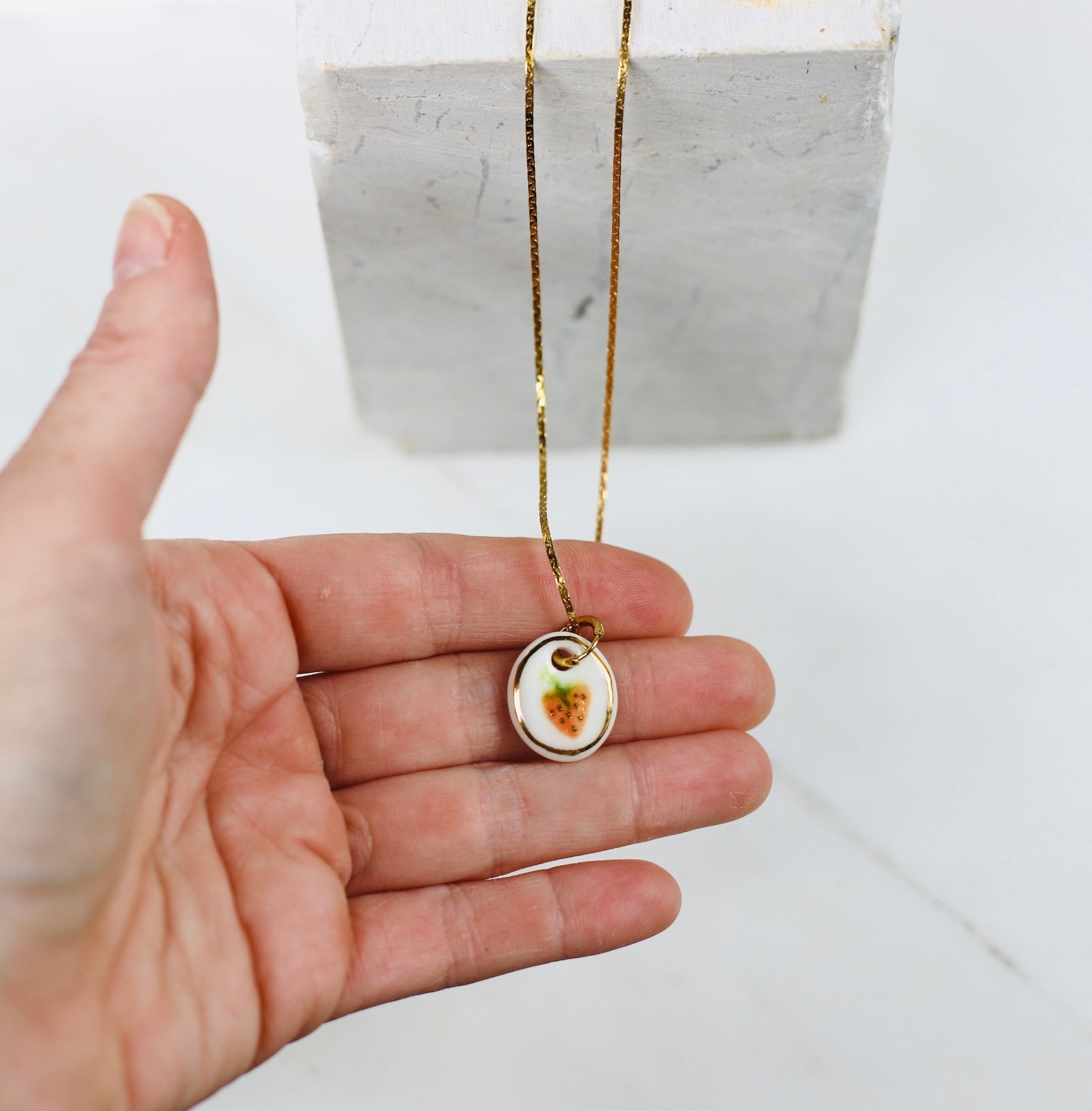 collier avec un médaillon de forme ovale en porcelaine blanche avec une fraise peinte à la main aux détails soignés avec des touches d'or liquide.